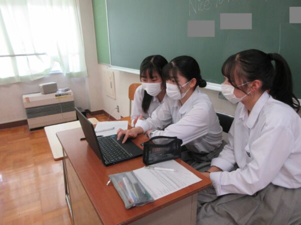 3人の生徒が黒板の前でラップトップPCを囲み、オンライン会議に参加している様子