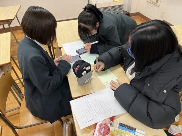 3人の生徒が机を囲んでグリーティングカードを書いている様子